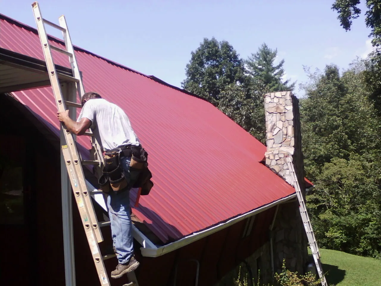 Roofer on a ladder beside a red metal roof and stone chimney — Soffit & Fascia Repair by Roofing Brothers in Chesterhill